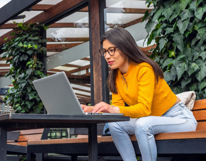 Young, Hispanic woman with indigenous features wearing a yellow sweater sitting on a bench outside while using her laptop